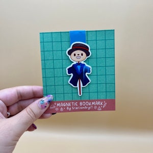 May include: A blue and white magnetic bookmark featuring a cartoon scarecrow with a top hat and a bow tie. The bookmark has the text "MAGNETIC BOOKMARK" and "By StationaryArt" on it.
