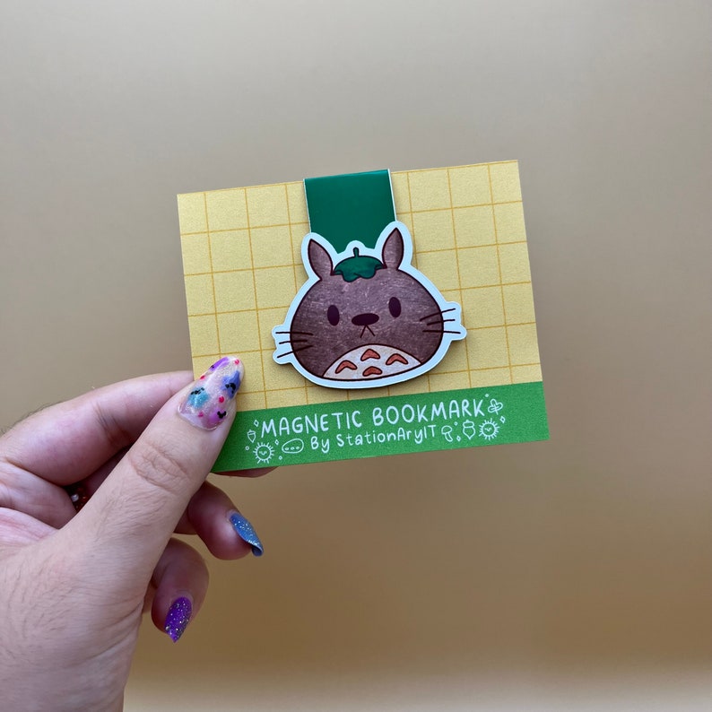 May include: A brown and white cartoon character with a green leaf on its head. The character is on a magnetic bookmark with a yellow and green grid background. The text "MAGNETIC BOOKMARK" and "By StationaryIT" is on the bookmark.