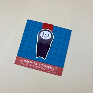 May include: A blue and red magnetic bookmark featuring a cartoon illustration of No Face from the movie Spirited Away. The bookmark has the text "Magnetic Bookmark - By StationaryArt" printed on the bottom.