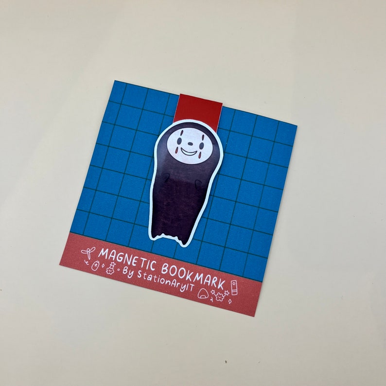 May include: A blue and red magnetic bookmark featuring a cartoon illustration of No Face from the movie Spirited Away. The bookmark has the text "Magnetic Bookmark - By StationaryArt" printed on the bottom.