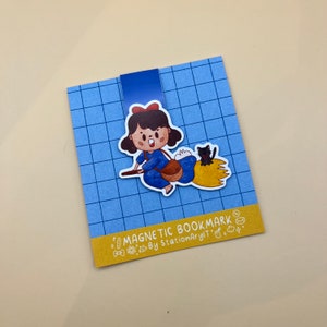 May include: A blue and yellow magnetic bookmark featuring a cartoon illustration of a girl riding a broom with a black cat. The bookmark has the text "MAGNETIC BOOKMARK by StationaryIT" on the bottom.