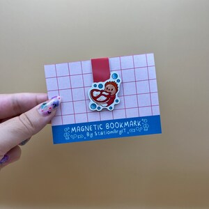 May include: A red and white magnetic bookmark with a cartoon illustration of a person in a red and white swimsuit. The bookmark is on a pink and white grid background. The text "MAGNETIC BOOKMARK" and "By StationaryIt." is printed on the bookmark.
