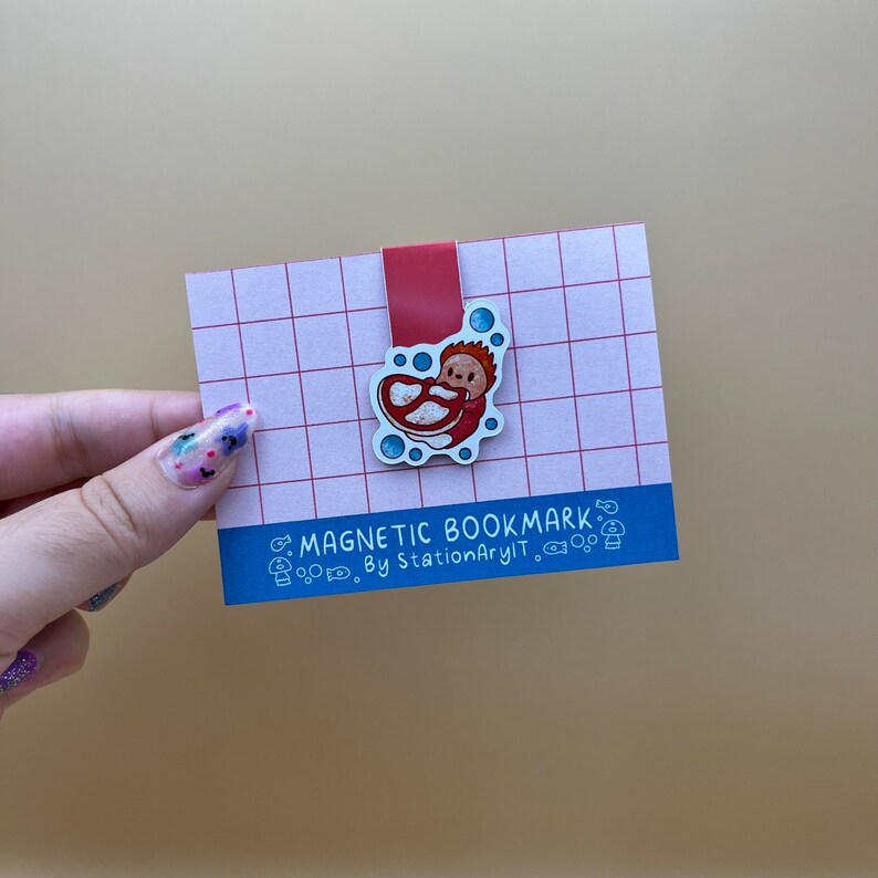 May include: A red and white magnetic bookmark with a cartoon illustration of a person in a red and white swimsuit. The bookmark is on a pink and white grid background. The text "MAGNETIC BOOKMARK" and "By StationaryIt." is printed on the bookmark.