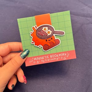 May include: A red and orange cartoon character shaped like a sausage with a happy face holding a frying pan with bacon and eggs. The character is on a green background with a white border. The text "MAGNETIC BOOKMARK" and "By StationaryArt" is on the bottom of the card.