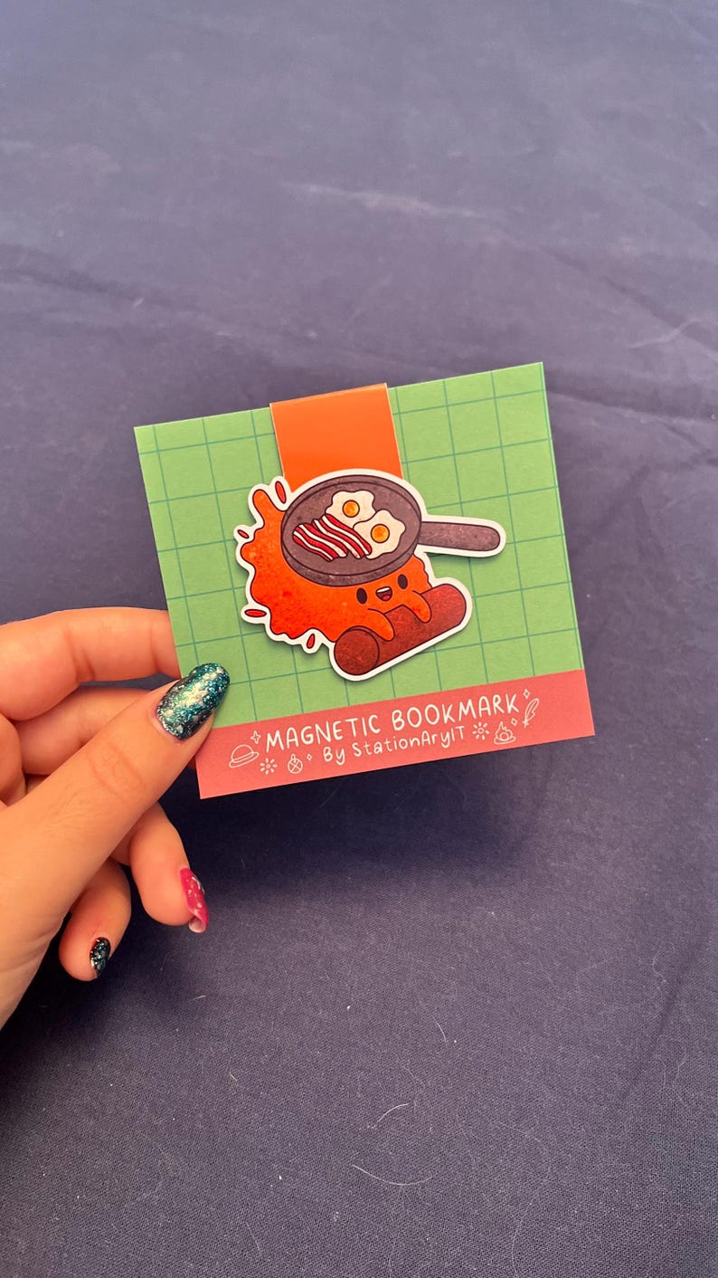 May include: A red and orange cartoon character shaped like a sausage with a happy face holding a frying pan with bacon and eggs. The character is on a green background with a white border. The text "MAGNETIC BOOKMARK" and "By StationaryArt" is on the bottom of the card.