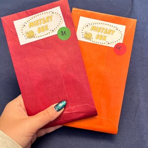 Può includere: Due scatole misteriose, una rossa e una arancione, con etichette che recitano "Mystery Box" e prezzi di 8 € e 6 €.