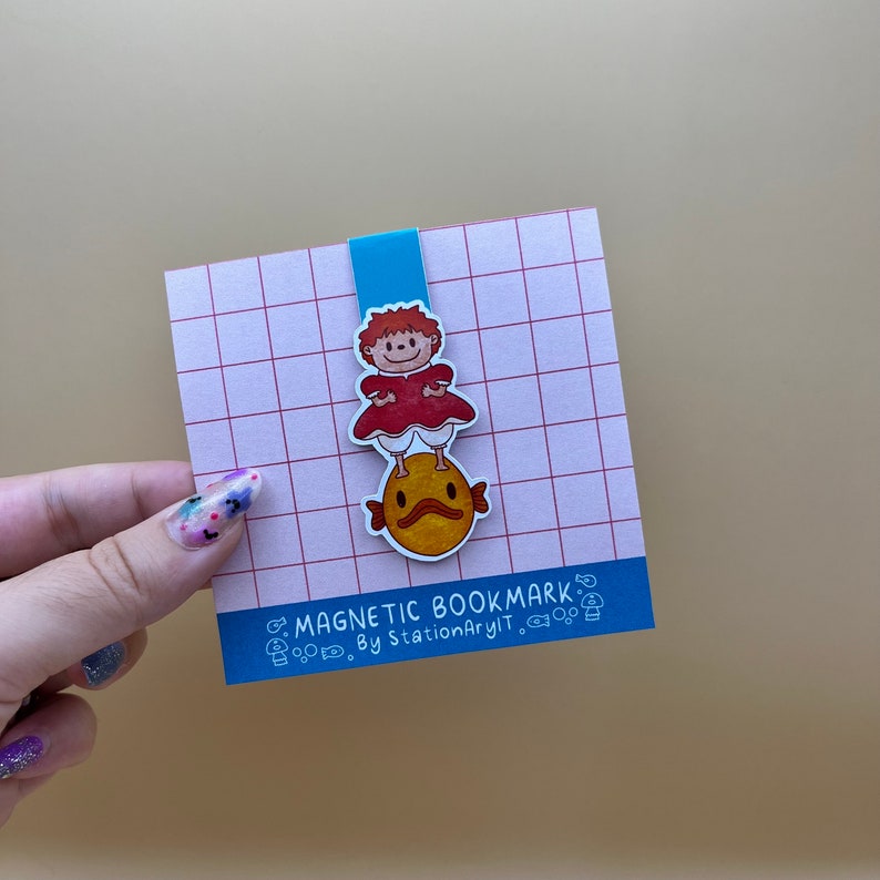 May include: A blue and white magnetic bookmark featuring a cartoon character in a red dress standing on a yellow fish. The bookmark has the text "MAGNETIC BOOKMARK. By StationaryIt." printed on it.