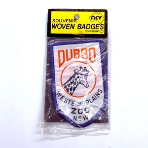 May include: A souvenir woven badge in a plastic sleeve. The badge features the word "Dubbo" in orange, a giraffe illustration, and the text "Western Plains Zoo NSW". The badge has a blue border and a yellow header.