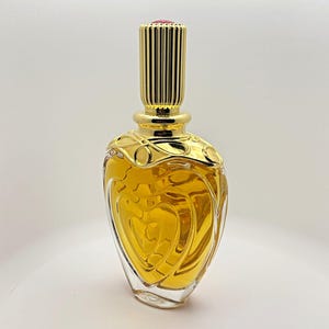May include: A clear glass perfume bottle with a heart-shaped design filled with amber-colored liquid. The bottle features a gold-toned metal collar with decorative swirling patterns and a tall, cylindrical, ribbed gold-toned metal cap.