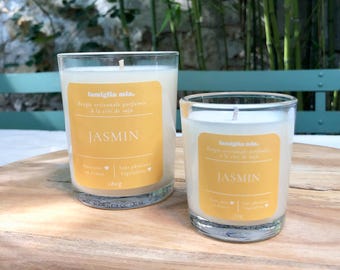 May include: Two clear glass candles with white wax and yellow labels that read "JASMIN". The candles are on a wooden surface. The larger candle has a weight of 180g.