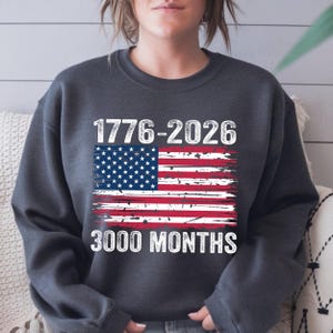 May include: Dark grey sweatshirt featuring a distressed American flag design. The graphic includes the years "1776-2026" and the text "3000 MONTHS" in white. A casual style sweatshirt.