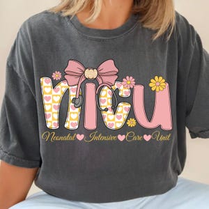 Peut inclure: Un t-shirt gris anthracite avec un motif représentant le texte NICU en grandes lettres roses et jaunes à motifs de cœurs. Un nœud rose et un stéthoscope sont placés au-dessus des lettres, accompagnés de petites fleurs jaunes et roses.