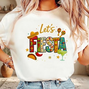 Può includere: T-shirt bianco con la scritta colorata "Let's Fiesta". Il design include un sombrero, maracas, un margarita, un taco e un peperoncino, il tutto in uno stile vivace e festoso.