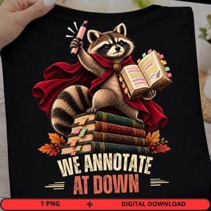 May include: A black t-shirt featuring a graphic of a raccoon wearing a red cape, holding a pink highlighter, and standing on a stack of books while reading an open book. The text reads WE ANNOTATE AT DOWN, surrounded by autumn leaves and colorful tabs.