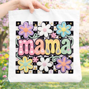 Peut inclure: Sac fourre-tout blanc avec un motif floral coloré et le mot "MAMA". Le motif comprend des fleurs aux couleurs pastel, un fond à carreaux et des étoiles. Le sac est tenu par une main.