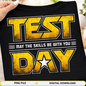 Peut inclure: T-shirt noir avec l'inscription "TEST DAY" en lettres jaunes et blanches. La phrase "MAY THE SKILLS BE WITH YOU" est centrée entre les mots. Une étoile blanche est dans la lettre "A".