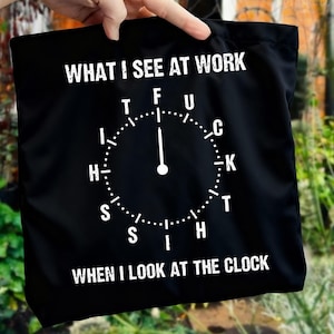 May include: Black tote bag with white text: "WHAT I SEE AT WORK" above a clock face with expletives instead of numbers. The text "WHEN I LOOK AT THE CLOCK" is below the clock face. The bag is held up by a hand.