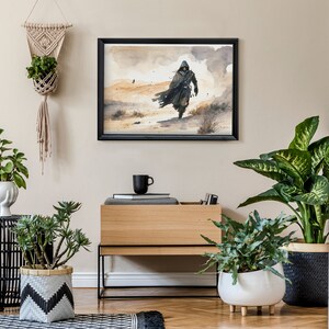 Puede incluir: Un interior de sala de estar con una pintura de acuarela enmarcada en negro que representa una figura encapuchada caminando en un paisaje des&eacute;rtico. La pintura est&aacute; colgada en una pared beige claro sobre un gabinete de madera con un marco de metal negro. Hay varias plantas en macetas en la habitaci&oacute;n, incluida una planta en una maceta colgante de macram&eacute;.