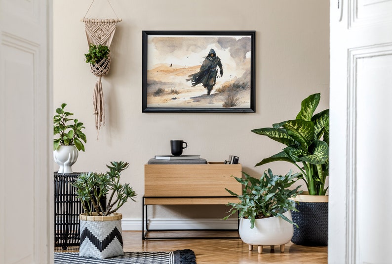 Puede incluir: Un interior de sala de estar con una pintura de acuarela enmarcada en negro que representa una figura encapuchada caminando en un paisaje des&eacute;rtico. La pintura est&aacute; colgada en una pared beige claro sobre un gabinete de madera con un marco de metal negro. Hay varias plantas en macetas en la habitaci&oacute;n, incluida una planta en una maceta colgante de macram&eacute;.