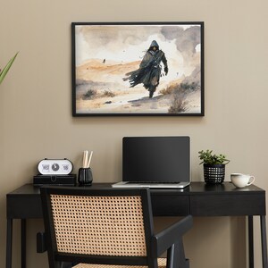Puede incluir: Una pintura al agua enmarcada de una persona con una capucha caminando por un paisaje des&eacute;rtico. La pintura est&aacute; colgada en una pared sobre un escritorio negro con una computadora port&aacute;til, una planta y una taza de caf&eacute;.