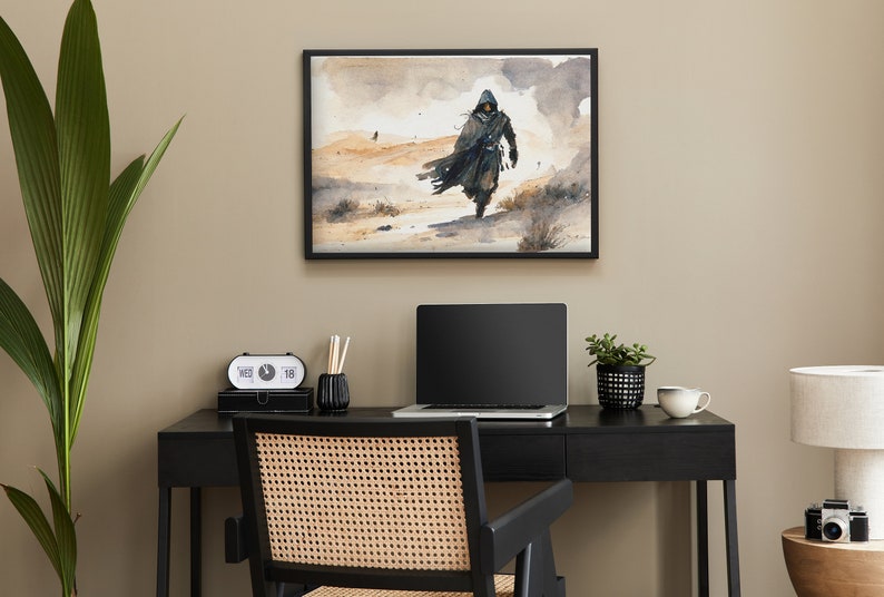 Puede incluir: Una pintura al agua enmarcada de una persona con una capucha caminando por un paisaje des&eacute;rtico. La pintura est&aacute; colgada en una pared sobre un escritorio negro con una computadora port&aacute;til, una planta y una taza de caf&eacute;.