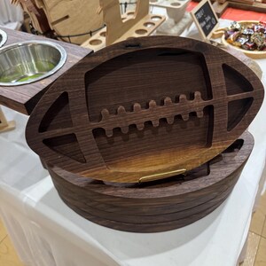 Puede incluir: Una bandeja de servir de madera con forma de balón de fútbol con un asa de metal dorado decorativa. La bandeja está hecha de madera de nogal oscuro y tiene un diseño tallado en el interior.
