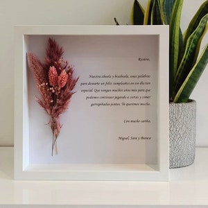 Puede incluir: Un marco blanco con un arreglo de flores secas en su interior. El marco tiene un mensaje escrito a mano que dice "Rosário, Nuestra abuela y bisabuela, unas palabras para desearte un feliz cumpleaños en ese día tan especial. Que vengan muchos años más para que podamos continuar jugando a cartas y comer garrapiñadas juntos. Te queremos mucho. Con mucho cariño, Miguel, Sara y Bianca"