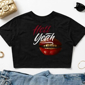 以下が含まれることがあります： 黒の半袖クロップトップ。赤い文字と白い文字で「Hell Yeah」と書かれ、その下に金色のグリルを装着した光沢のある赤い唇のグラフィックが描かれています。シャツはゴールドのハンドバッグ、サングラス、デニムと一緒に配置されています。