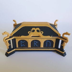 Puede incluir: Una caja de madera negra con detalles dorados y asas con cabeza de dragón. La caja tiene tres secciones arqueadas con runas azules que dicen "KRN".