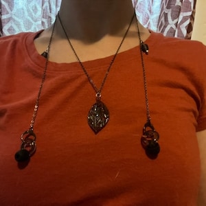 May include: A dark metal necklace with a leaf pendant and two dangling chains. Each chain ends with a black bead and a decorative metal element. The necklace is worn over a red t-shirt.
