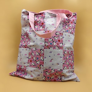 Könnte beinhalten: Eine rosa und graue Patchwork-Tragetasche mit einem Einhorn-Druck. Die Tasche hat einen rosa Griff und ist aus Baumwollstoff.