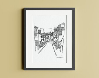 May include: Black and white line drawing of a narrow street with buildings on either side. The street is lined with trees and the buildings have windows and doors. The drawing is framed in a black frame.