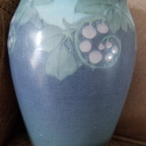 May include: A blue ceramic vase with a crackle glaze. The vase features a design of green leaves and pink berries around the rim. The vase has a rounded shape and a smooth, glossy finish.