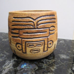 Op de afbeelding: Een bruine keramische vaas met een gegraveerd ontwerp van een gezicht. De vaas heeft een ronde vorm en een glad, getextureerd oppervlak.