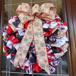May include: A festive wreath with a large bow, featuring red, white, and blue ribbons. The bow has a tan background with red and blue firework designs. The wreath is made of layered ribbons in various patterns and colors.