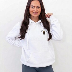 May include: White hoodie with a front pocket and drawstring. The hoodie has the text "SPLIT THE G" with a graphic of a pint of stout. The model is wearing gray leggings.