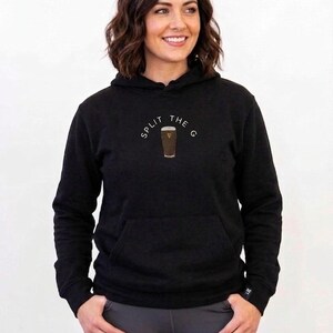 May include: Black hoodie with a front pocket and drawstring hood. The hoodie features the text "SPLIT THE G" in a semi-circle above a graphic of a pint of stout. The model is wearing gray leggings.