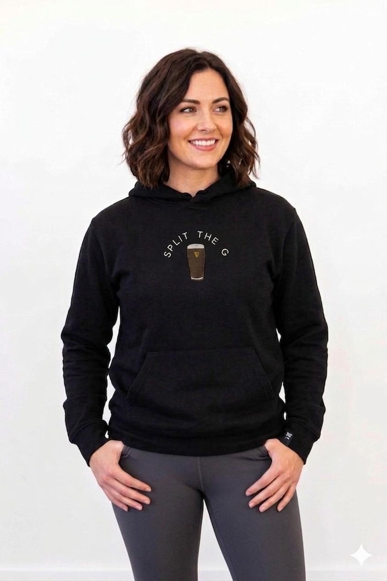 May include: Black hoodie with a front pocket and drawstring hood. The hoodie features the text "SPLIT THE G" in a semi-circle above a graphic of a pint of stout. The model is wearing gray leggings.