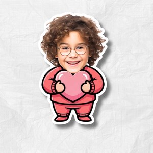 May include: A sticker featuring a cartoon illustration of a child with curly brown hair and glasses, holding a pink heart. The child is wearing a pink outfit. The sticker has a white border and is set against a textured white background.