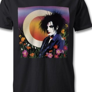 Pode incluir: T-shirt preto com estampa gráfica. O gráfico mostra uma pessoa com cabelo escuro e maquiagem, em um fundo de um grande círculo claro e flores coloridas. A pessoa está vestindo uma camisa azul.