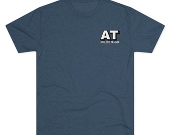 Può includere: Una maglietta blu a maniche corte con le lettere "AT" in bianco e nero, con le parole "ATHLETIC TRAINER" sotto. La maglietta ha un girocollo ed è realizzata in un materiale morbido.