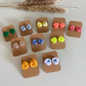 Puede incluir: Una colección de pendientes de ganchillo hechos a mano en varios colores, incluyendo verde, azul, amarillo, rosa y blanco. Cada par está unido a una pequeña tarjeta de presentación de cartón marrón. Los pendientes presentan un diseño anudado y texturizado.