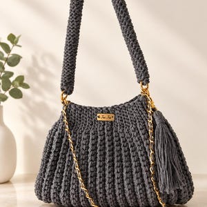 Puede incluir: Un bolso de ganchillo gris oscuro con un asa gruesa y texturizada y una correa de cadena dorada. El bolso presenta un patrón de canalé vertical, una borla gris a juego y una pequeña etiqueta metálica rectangular de color dorado con el texto Fini Fini.