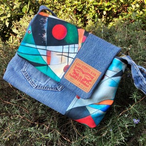 Può includere: Una borsa in denim blu con una stampa astratta colorata e una toppa Levi's. La borsa ha una tracolla lunga e una chiusura a lembo.