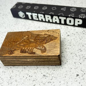 May include: A wooden box with a bear design etched on the top. The box is brown and rectangular. A black box with the word "TERRATOP" in white is in the background. The box has a dice pattern.