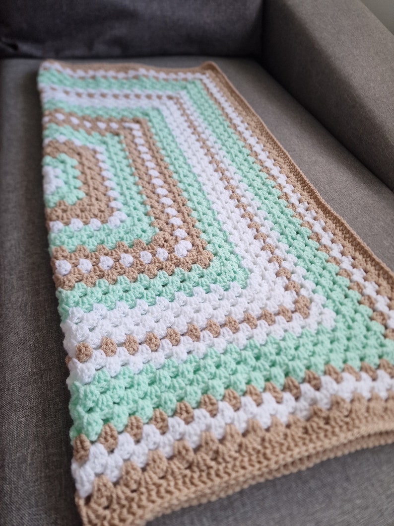 May include: A crocheted baby blanket with a green, white, and brown striped pattern. The blanket is made with a textured stitch that creates a raised, bumpy surface.