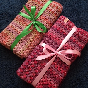 May include: Two crocheted blankets, one pink and one with shades of pink, orange, and brown, are tied with ribbons. The pink blanket has a pink ribbon, and the other blanket has a green ribbon.