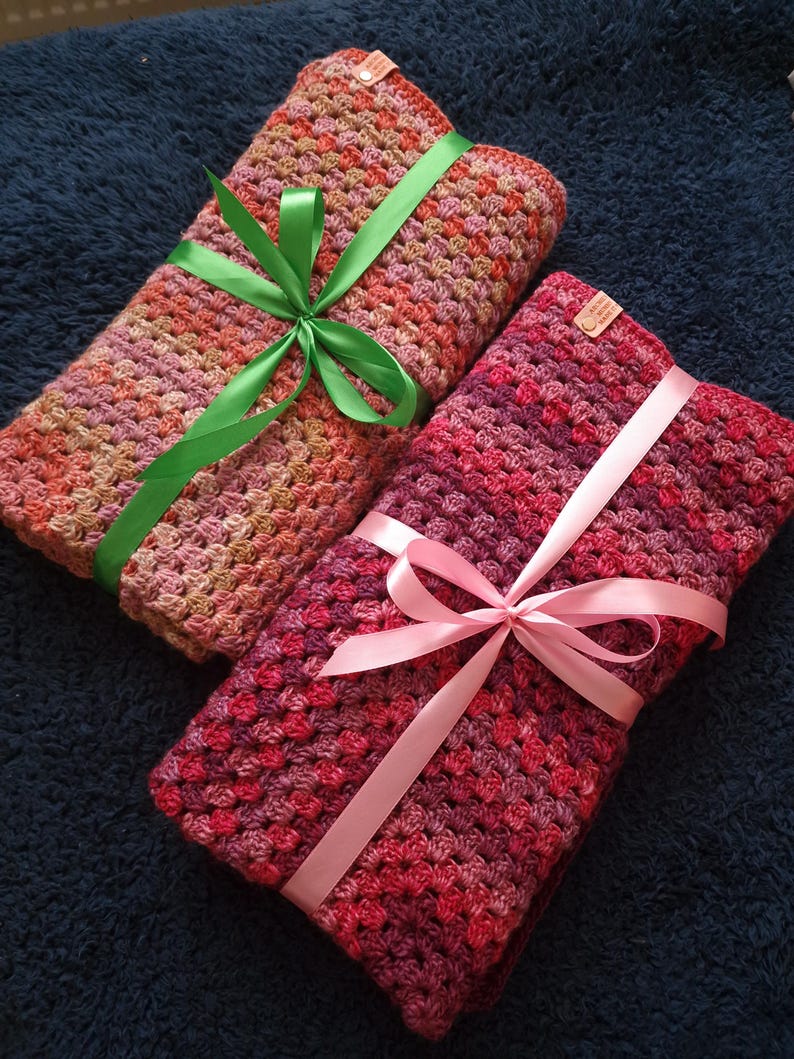May include: Two crocheted blankets, one pink and one with shades of pink, orange, and brown, are tied with ribbons. The pink blanket has a pink ribbon, and the other blanket has a green ribbon.