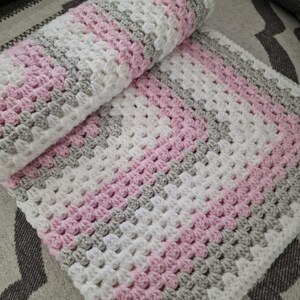 May include: A crocheted baby blanket with pink, white, and gray stripes. The blanket is made with a textured stitch and is folded in half.