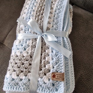 May include: A blue, white, and gray crocheted baby blanket tied with a light blue ribbon. A brown leather tag with the text "Archie's Mummy Made It" is attached to the blanket.